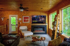 A cozy cabin interior with a fireplace, a television mounted above it, and a large window offering a view of the forest.