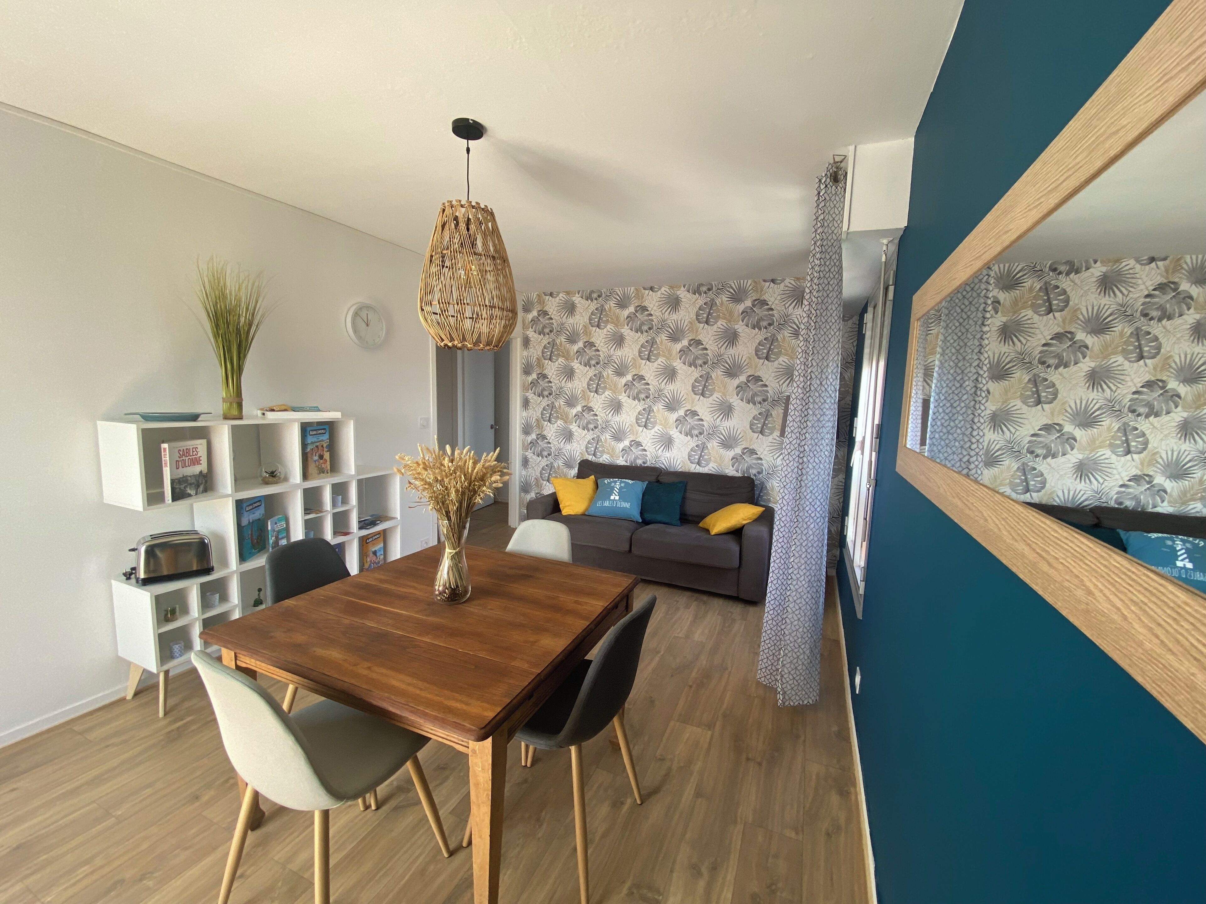 A modern living room with a wooden dining table, a sofa with patterned cushions, and a decorative hanging light fixture.