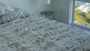 Ein Schlafzimmer mit Bett, Nachttisch, Lampe und Blick auf eine Stadtlandschaft durch ein großes Fenster.