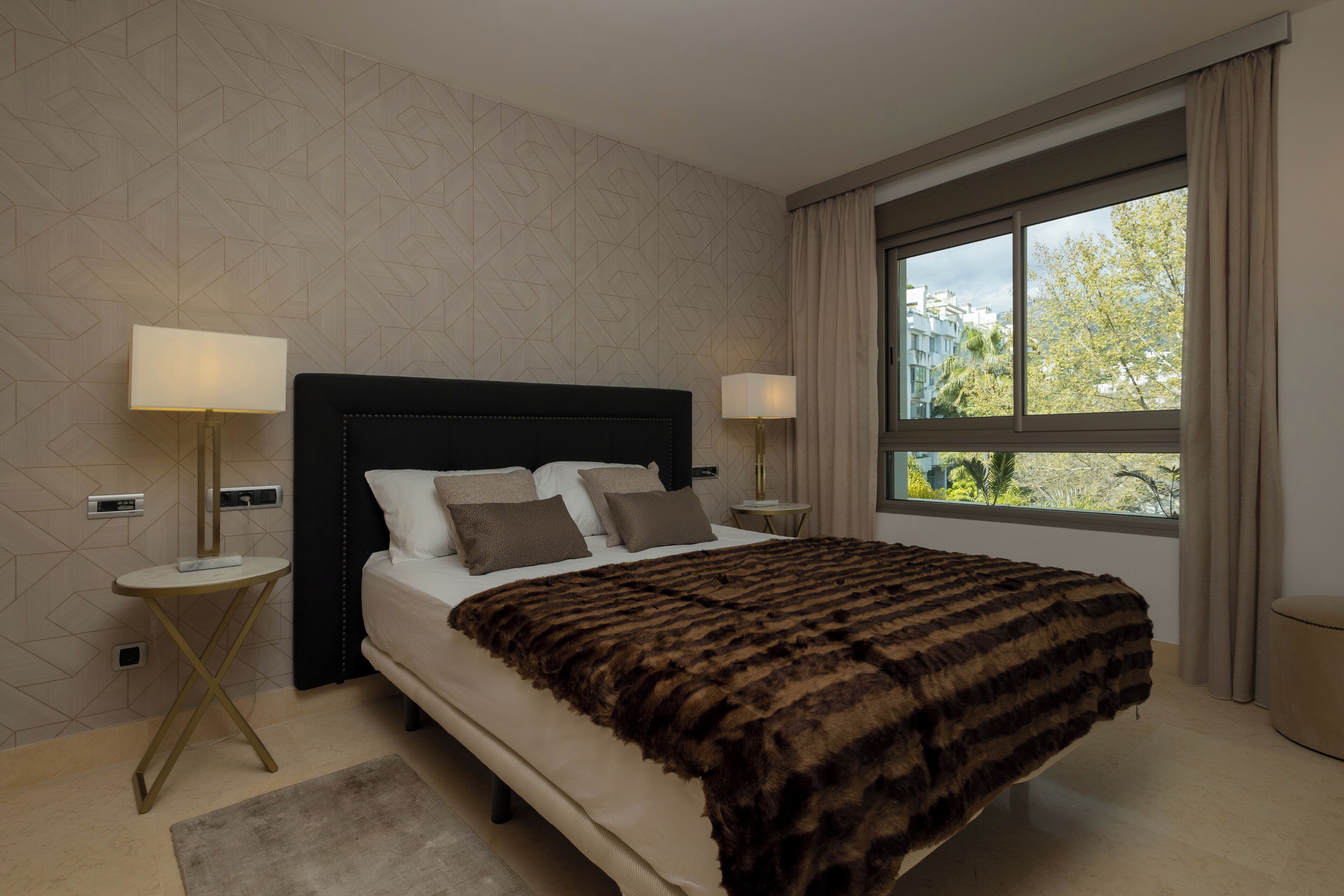 Een moderne hotelkamer met een groot bed, twee nachtkastjes met lampen, en een uitzicht op gebouwen door een groot raam, met een kamer aan de muur.