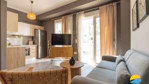 A modern living room with a sofa, a dining area with a table, and a kitchen with a refrigerator and a TV. - 2BD Modern Home Steps from Pasalimani (Pireas)