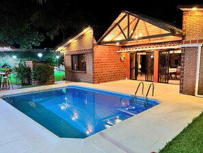 Piscina com deck ladrilhado, casa de tijolos com porta de vidro e estrutura de telhado de palha.