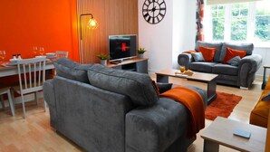 Ett modernt vardagsrum med en grå soffa, ett matbord med stolar, en tv och ett stort fönster med gardiner.