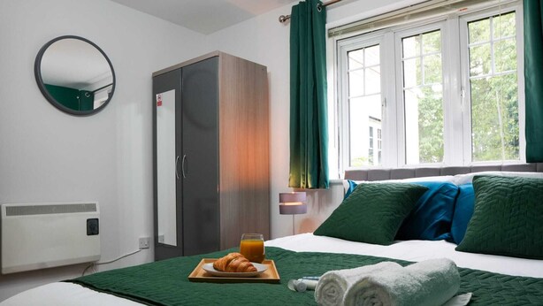 Ett hotellrum med en säng, grönt sänglinne, ett sängbord med en lampa, en garderob och ett fönster med gardiner.