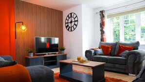 Ett modernt vardagsrum med en platt-TV, en soffa med orange kuddar, ett soffbord och ett stort fönster med gardiner.