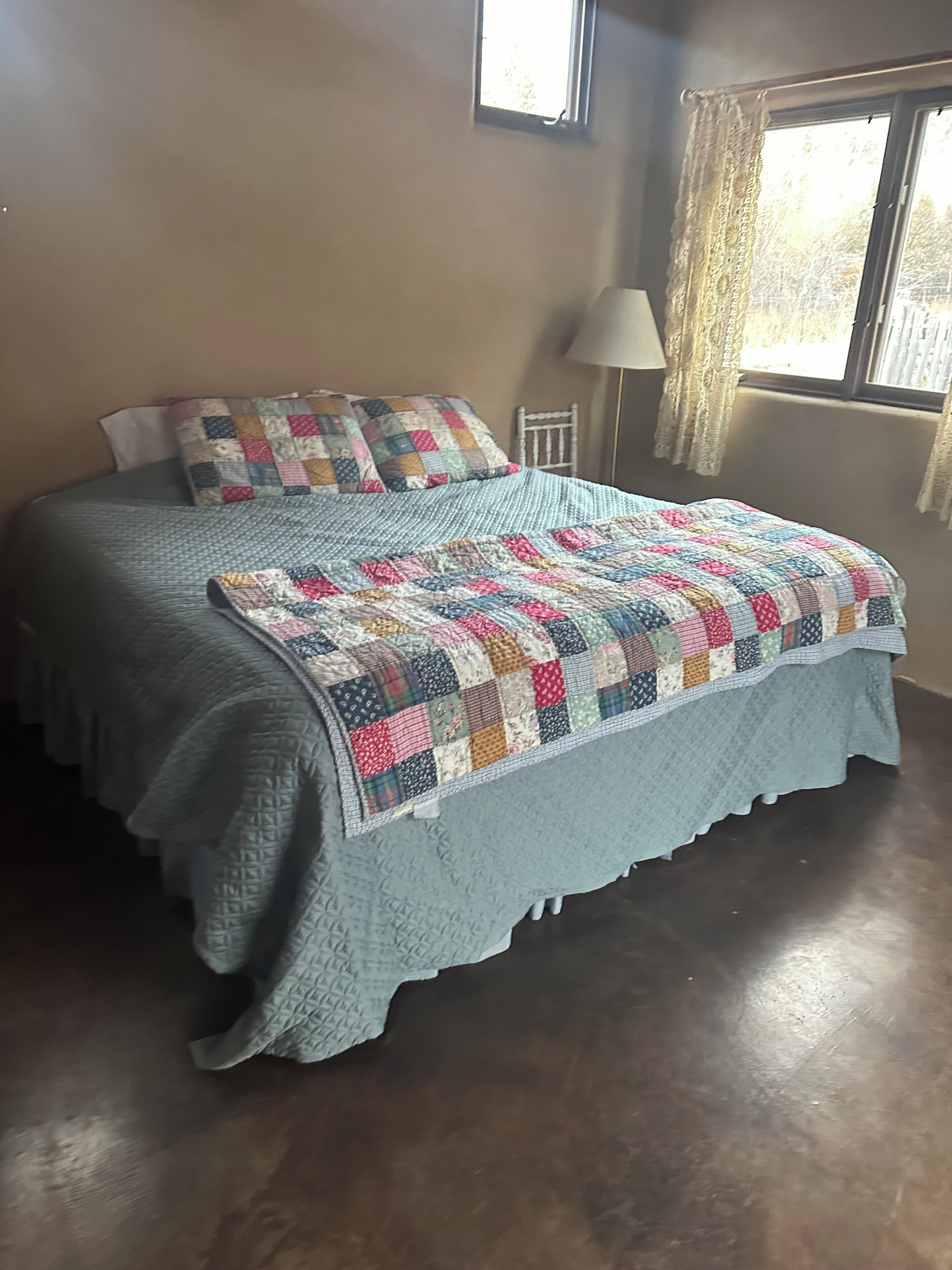 Una cama con colcha, una lámpara y una ventana con cortinas.
