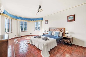 Un dormitorio con cama, mesitas de noche, ventana con cortinas, un cuadro en la pared y suelo de baldosas.