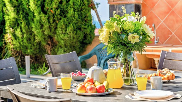 Ein Tisch reichhaltig mit Frühstück zubereitet, darunter Früchte, Gebäck und Getränke, im Freien mit Stühlen und einer dekorativen Vase.