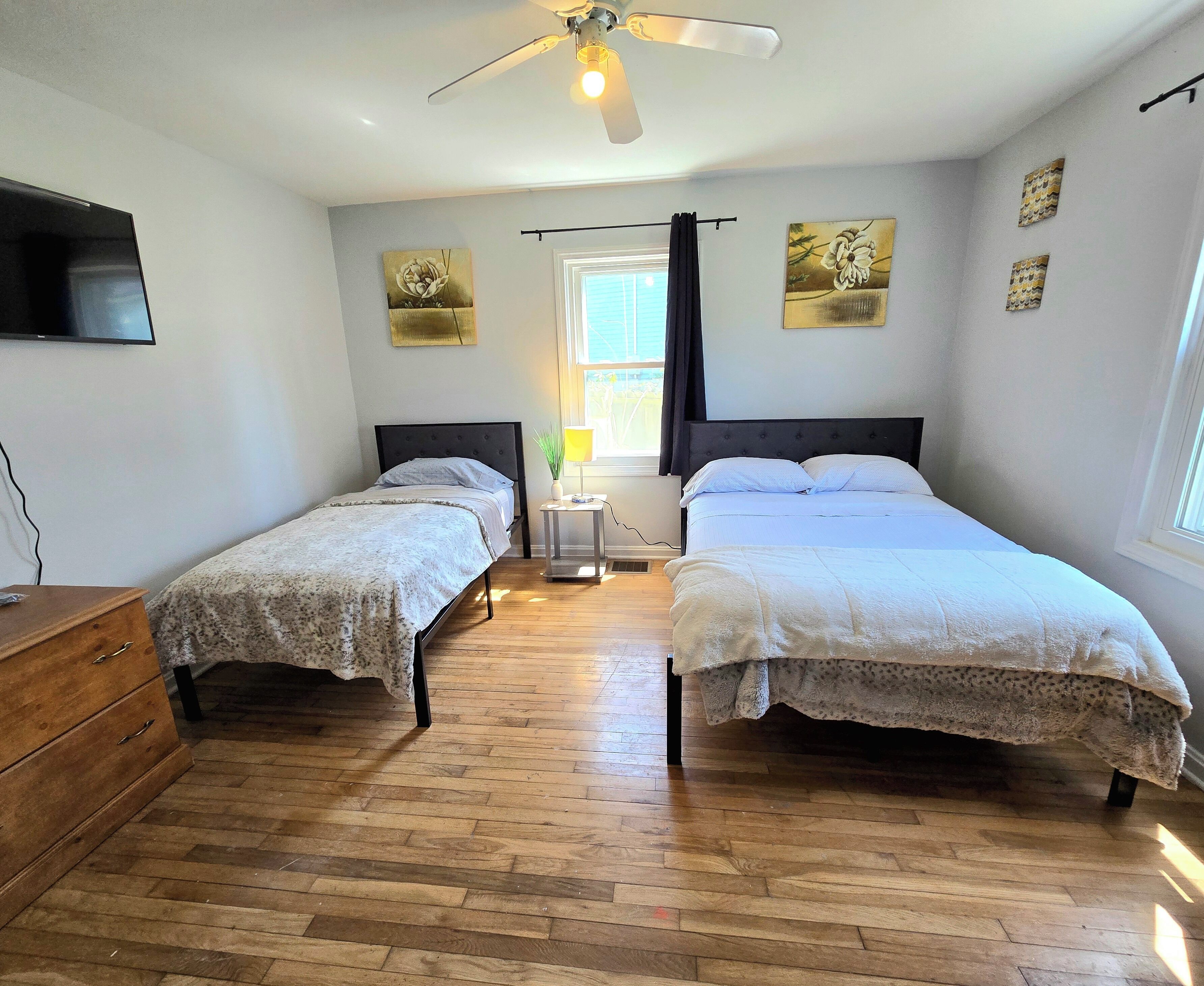 Une chambre avec deux lits, un ventilateur de plafond, une fenêtre avec des rideaux et des œuvres d’art accrochées aux murs.