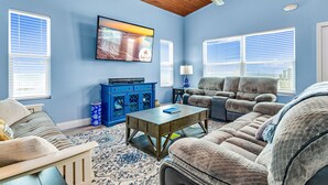 A living room with a blue wall, wooden ceiling, a flat-screen TV, a blue cabinet, a sofa, a coffee table, and a patterned rug. - Beach Blast: Gulf-Front 3bd/2ba on North Cape - Ocean Views From Every Room (Port St. Joe)