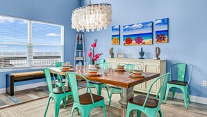 A dining area with a wooden table, teal chairs, and a chandelier. There are three paintings on the wall depicting beach scenes. - Beach Blast: Gulf-Front 3bd/2ba on North Cape - Ocean Views From Every Room (Port St. Joe)
