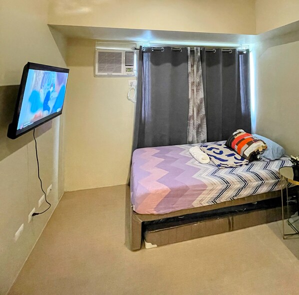 Un dormitorio pequeño con cama, televisor montado en la pared y ventana con cortinas.