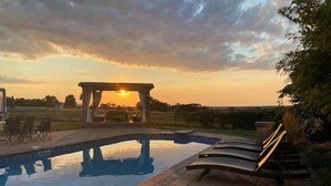 A swimming pool with lounge chairs, a pergola, and a sunset view.