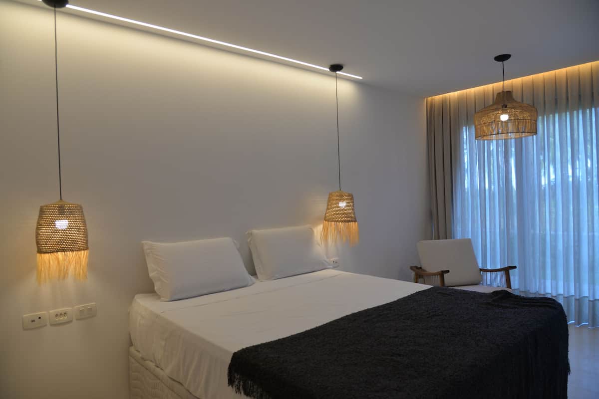 Een moderne slaapkamer met een groot bed, twee bedlampen, een stoel en een raam met dunne gordijnen.