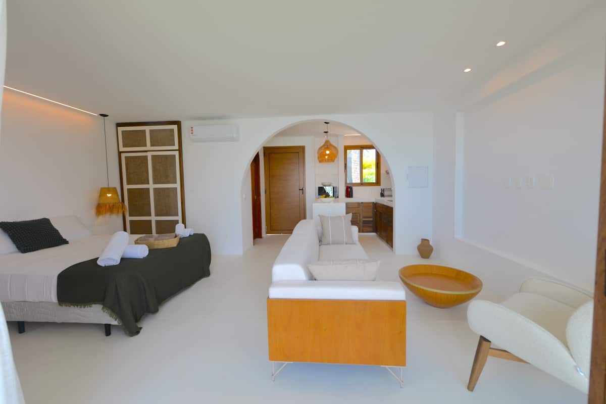 Een moderne woonkamer met een bank, een houten salontafel en een witte fauteuil. Een bed met donkere hoofdbord en een lamp bevindt zich aan de linkerkant. Een boogvormige doorgang leidt naar een keuken.