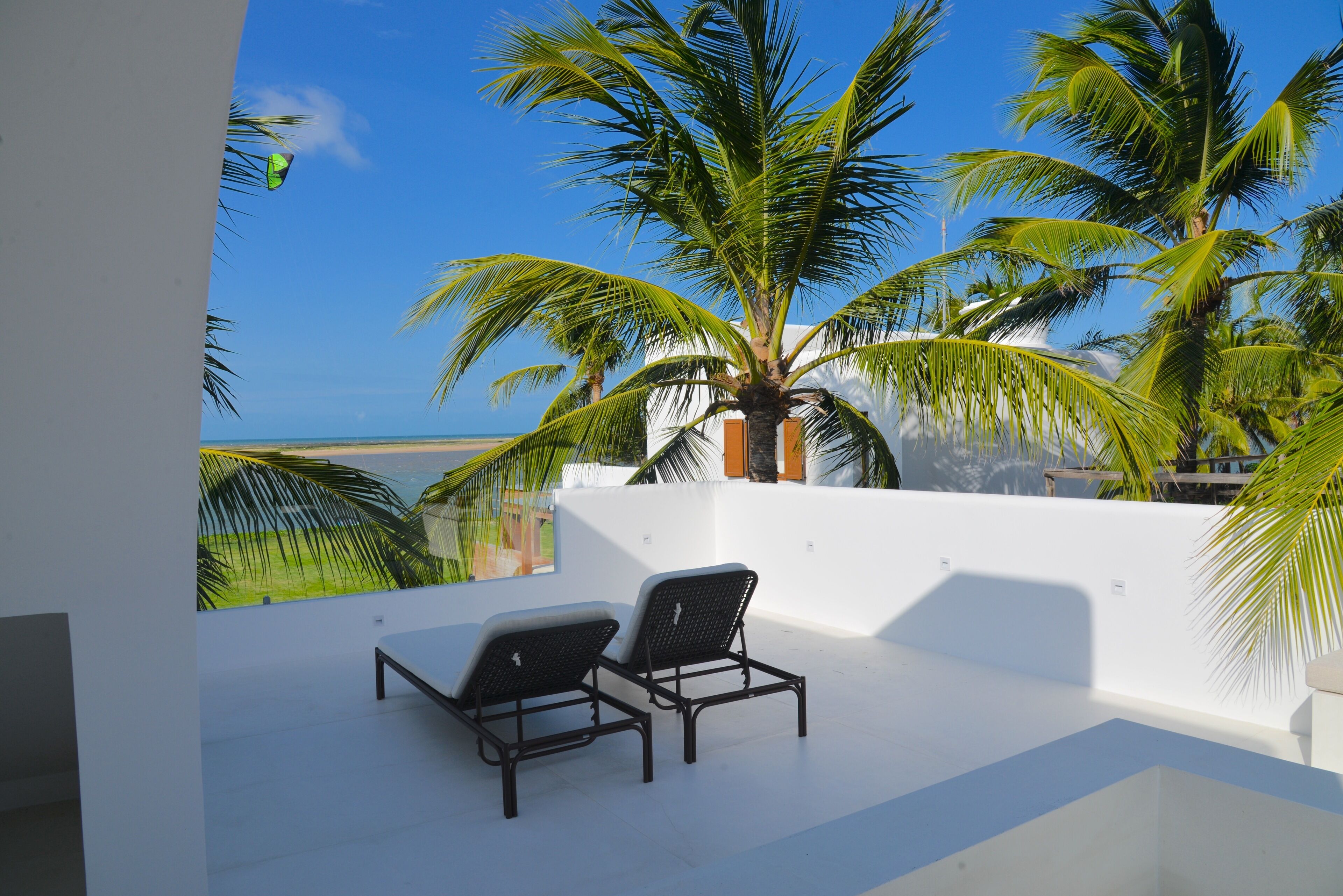 A balcony with two black lounge chairs, a white wall, and a view of the ocean and palm trees.