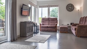 A living room with a grey sofa, a brown armchair, a TV on a cabinet, and a clock on the wall. - Perfect for families and couples alike, a short walk to the lovely sandy beach. (Hemsby)