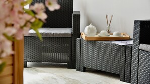 A wicker chair with a cushion, a small table with a tray, and a vase with reeds. - Alight Sauna and SPA Penthouse (Xewkija)