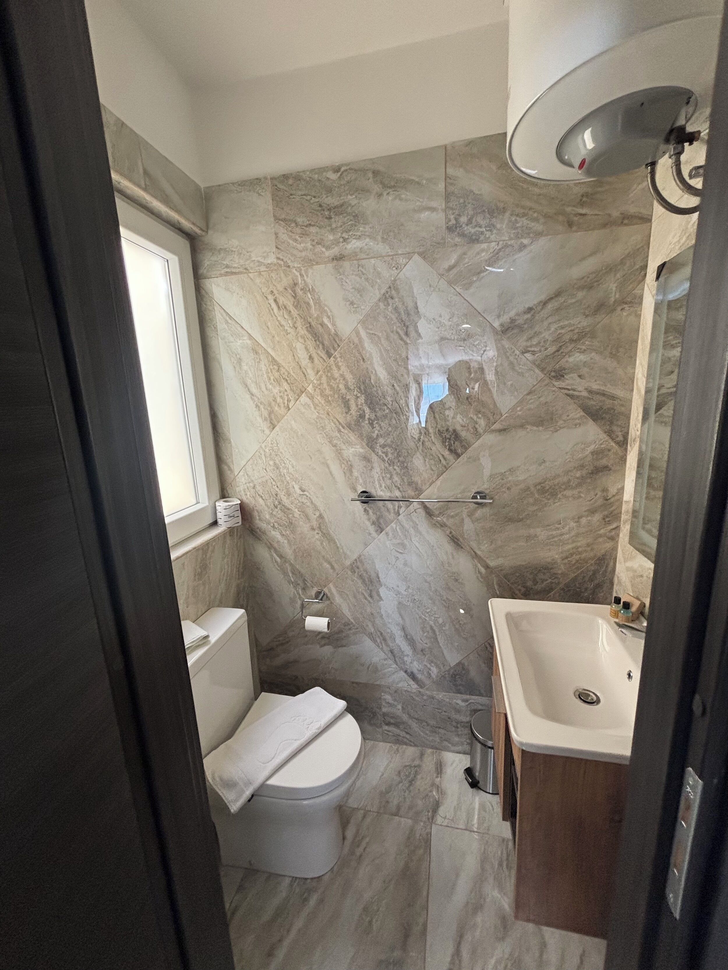 A bathroom with a toilet, shower, and sink, featuring marble tiles and a window.