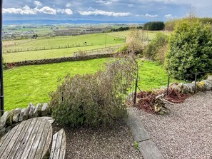 A rural landscape with a stone wall, a wooden table, and a view of green fields and distant hills.