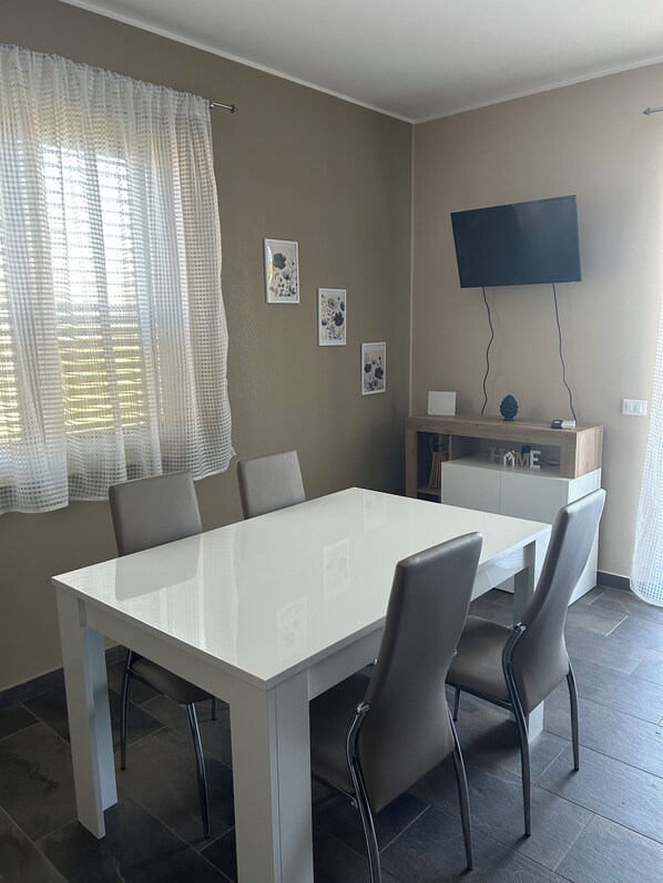 A modern dining area with a white table and chairs, a flat-screen TV mounted on the wall, and framed artwork on the wall. - Panorama d'oro (Sicilia)
