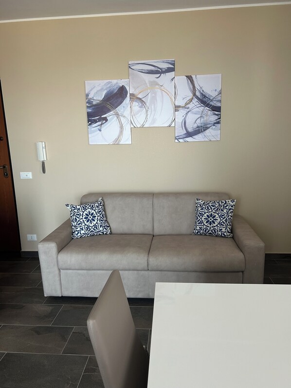 A neutral-toned living room with a beige sofa, two patterned throw pillows, and abstract wall art. - Panorama d'oro (Sicilia)