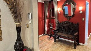 A room with a red wall, a wooden bench, a mirror, a clock, and a vase with dried plants. - Departamento Por Temporadas (La Paz)