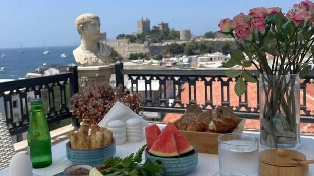 Une table avec de la nourriture et des boissons, une sculpture en buste, et une vue sur la mer et les bâtiments.