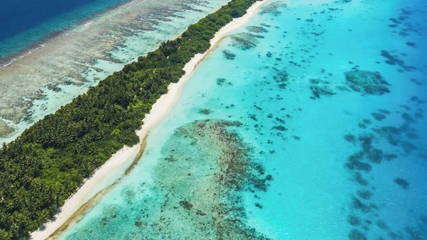 An aerial view of a coastal area with a long stretch of beach, clear turquoise waters, and dense green vegetation along the shoreline. - Azul Retreat (Dhigurah)