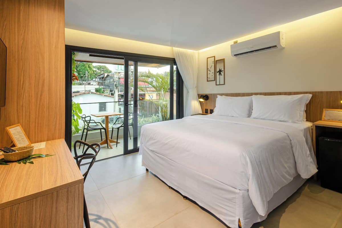 A modern hotel room with a large bed, a desk with a chair, a balcony with a table and chairs, and an air conditioning unit.