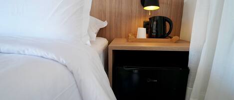 Habitación de hotel con cama, mesita de noche con lámpara, cafetera y lavavajillas negro.