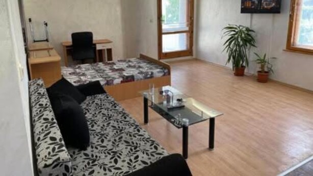 A small, well-lit room with a bed, a sofa, a small table, and a TV on the wall.