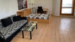 A small, furnished room with a sofa, a bed with a patterned bedspread, a small table, and a chair.