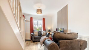 Un salon moderne avec un canapé, une table basse et une lampe. Un escalier se trouve à gauche, et une fenêtre est agrémentée de rideaux.