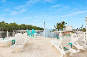 A rooftop terrace with white and turquoise lounge chairs, a pool, and a view of the ocean and buildings.