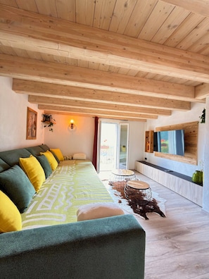 A room with a wooden ceiling, a sofa with cushions, a TV mounted on the wall, and a small table with chairs.