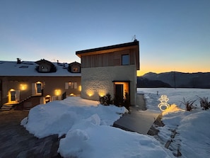 A modern two-story house with a snow-covered driveway and a mountain backdrop.