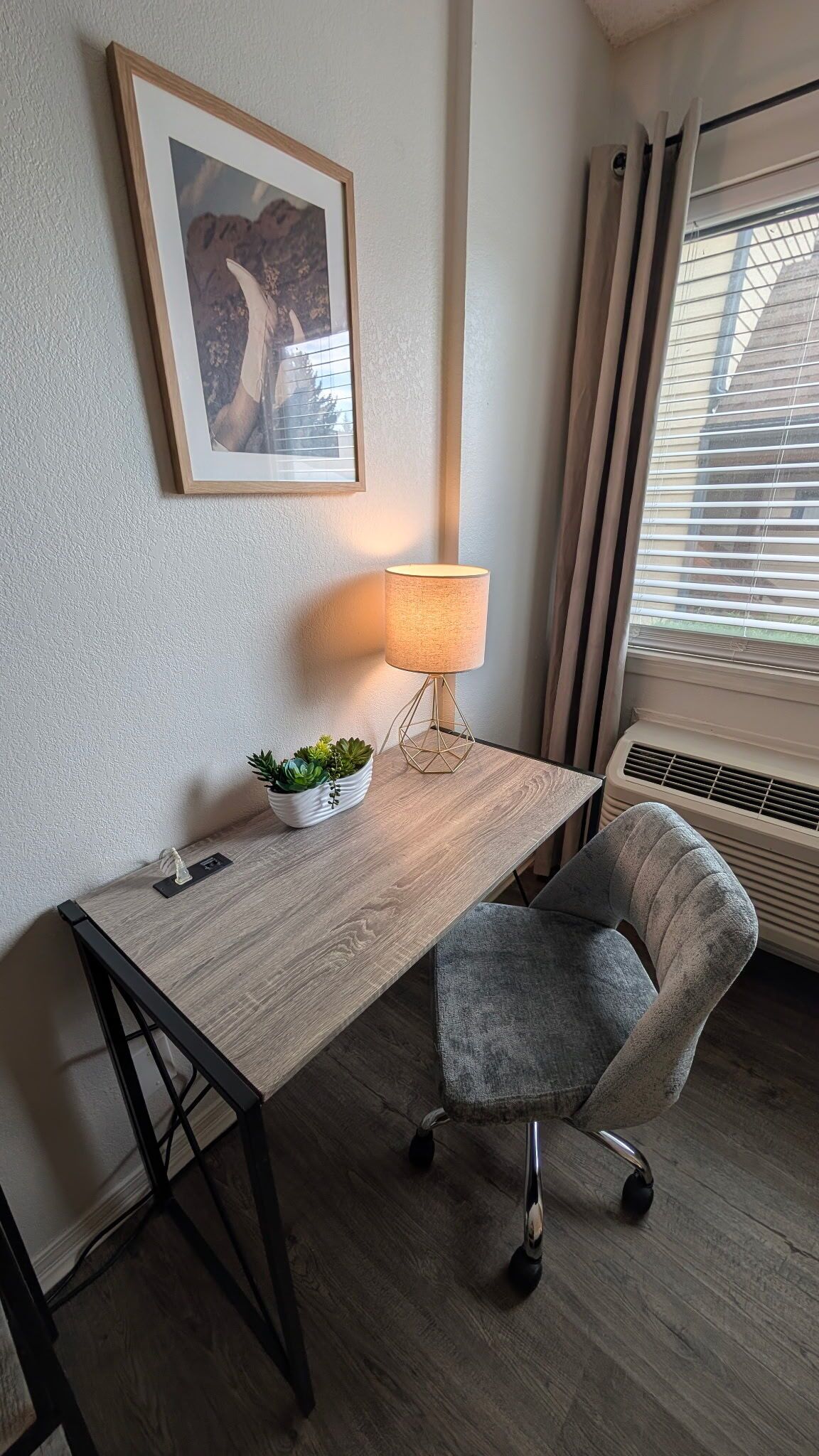 A room with a wooden desk, a grey office chair, a floor lamp, a framed picture, a potted plant, and a window with blinds.