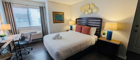 Una habitación de hotel con una cama, dos mesitas de noche con lámparas, un escritorio con una silla, un ventanal con cortinas y un adorno en la pared.