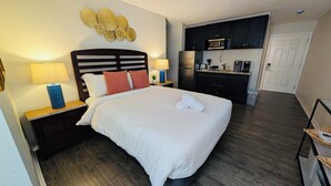 A hotel room with a bed, nightstands, a lamp, a wall decoration, a kitchenette, and a door.