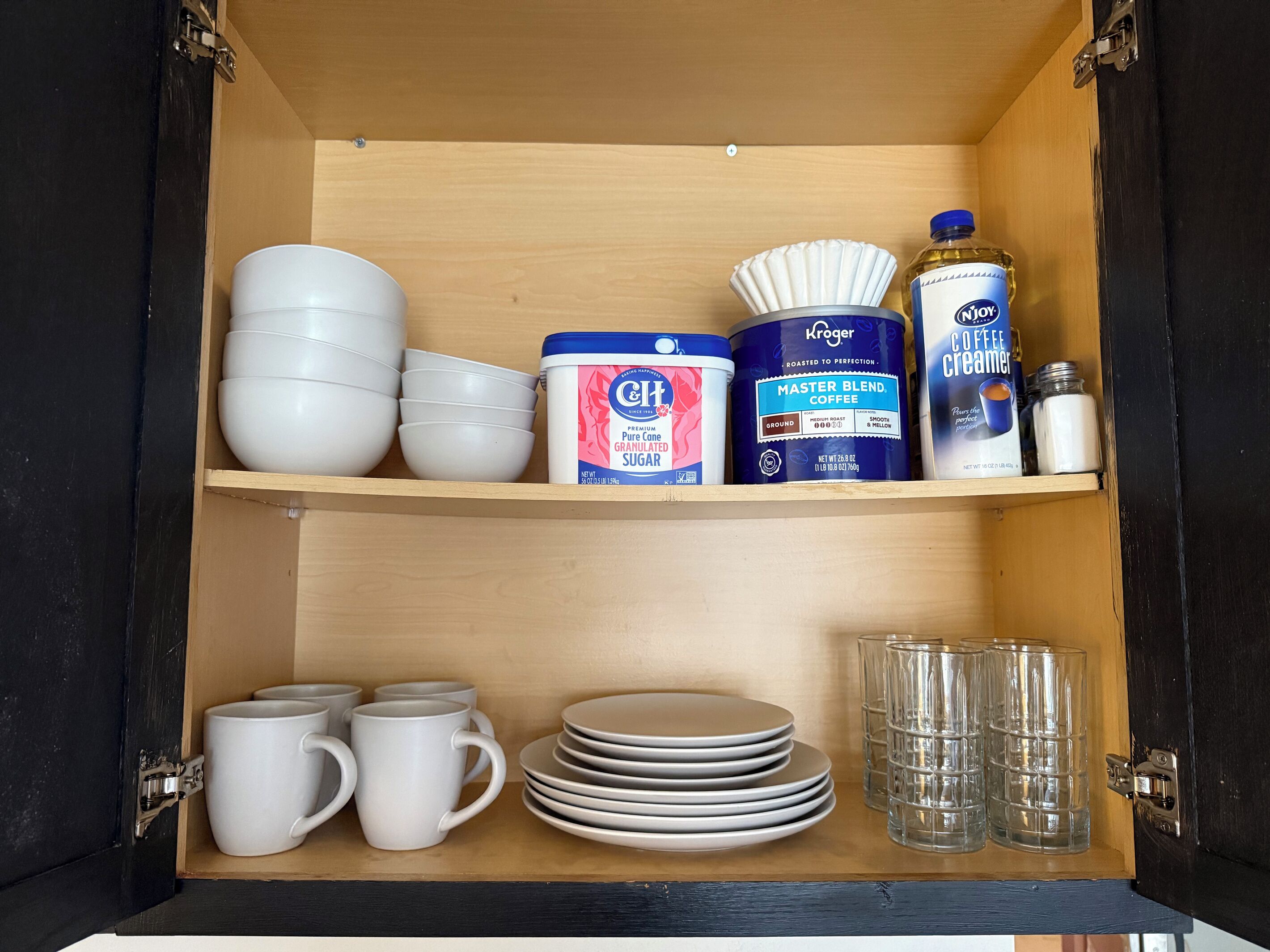 A kitchen cabinet with white bowls, a container of sugar, a coffee creamer, and a coffee grinder.
