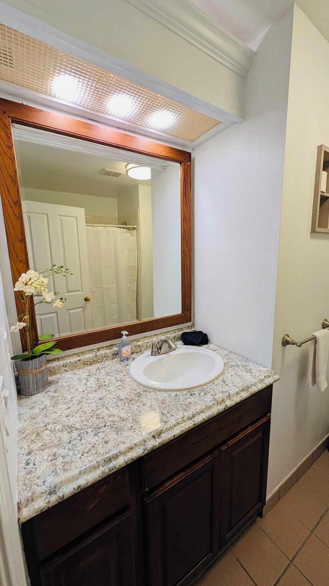 Un baño con encimera de granito, un lavabo blanco, un espejo con marco de madera y un perchero para toallas.