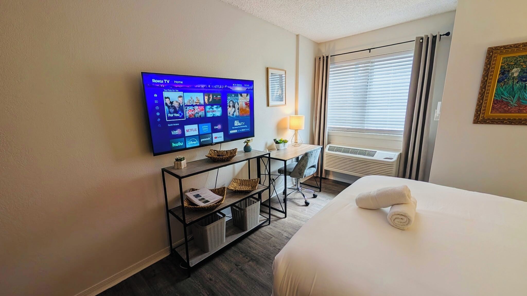 Ett hotellrum med en platt-TV som visar gränssnittet för en streamingtjänst, ett skrivbord med en lampa, en stol och en säng med en hoprullad handduk.