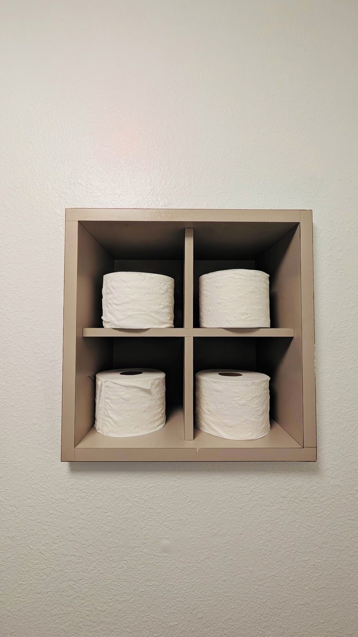 A wall-mounted storage unit with four compartments, each containing a roll of toilet paper.