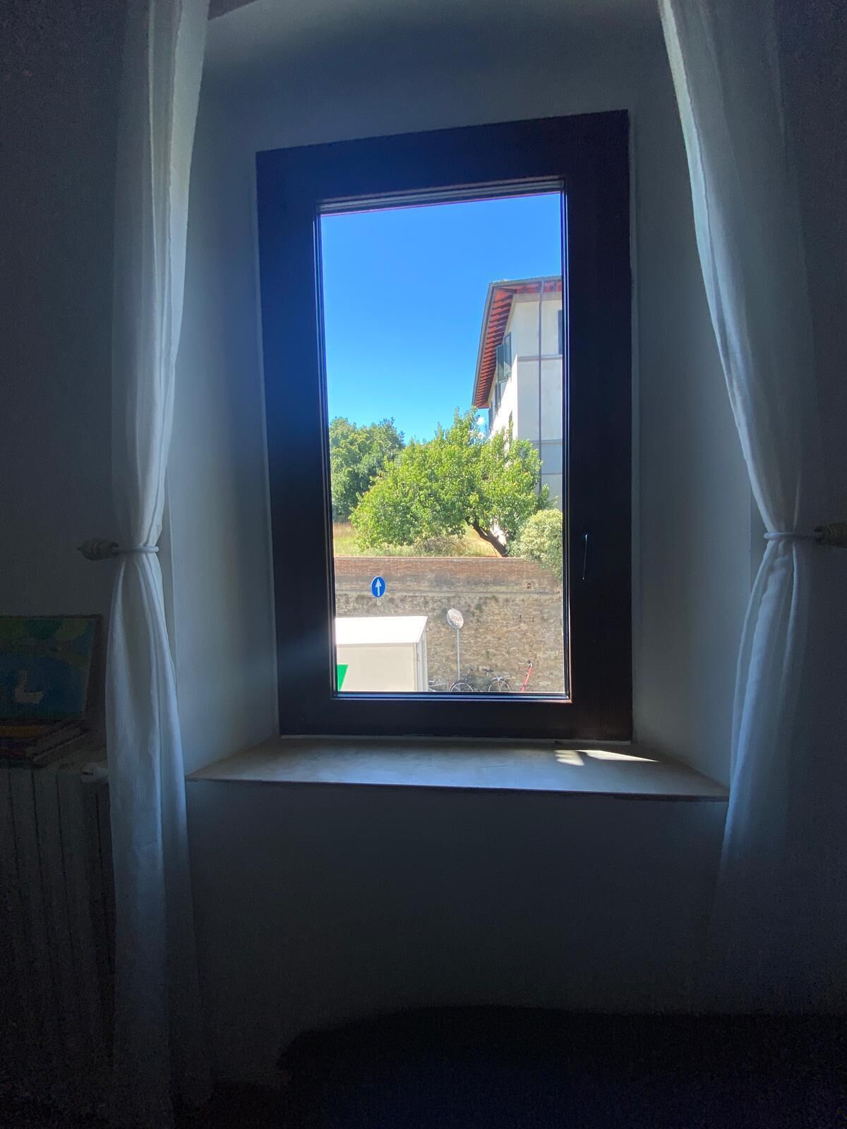A window with white curtains, a view of a building, and a clear blue sky.
