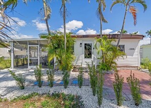 Una casa a un piano con facciata bianca, tetto rosso e il numero '12070' sul lato. Dispone di un portico coperto con mobili da esterno e un piccolo giardino con diverse piante e una palma.