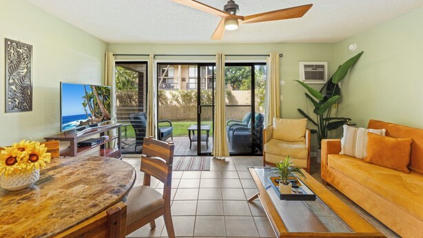 A living room with a ceiling fan, a sofa, a coffee table, a TV, and a dining area with a table and chairs. - Charming Tropical Kihei Gardens 2-Bed Suite (Kihei)
