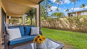 A covered outdoor seating area with a wicker sofa and a glass table, a vase with sunflowers, and a view of a well-maintained lawn and palm trees. - Charming Tropical Kihei Gardens 2-Bed Suite (Kihei)
