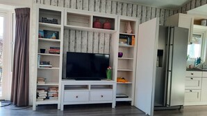 A living room with a TV on a white entertainment unit, a bookshelf, and a kitchen area with a fridge. - Luxury, space, comfort and nature for two: The Veluwe Happy Together! (Epe)