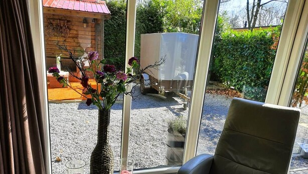 A room with a large window, a chair, a vase with flowers, and a covered vehicle outside. - Luxury, space, comfort and nature for two: The Veluwe Happy Together! (Epe)